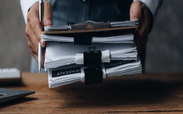 Two hands holding a stack of documents, with a pen also visible, in a professional office environment.